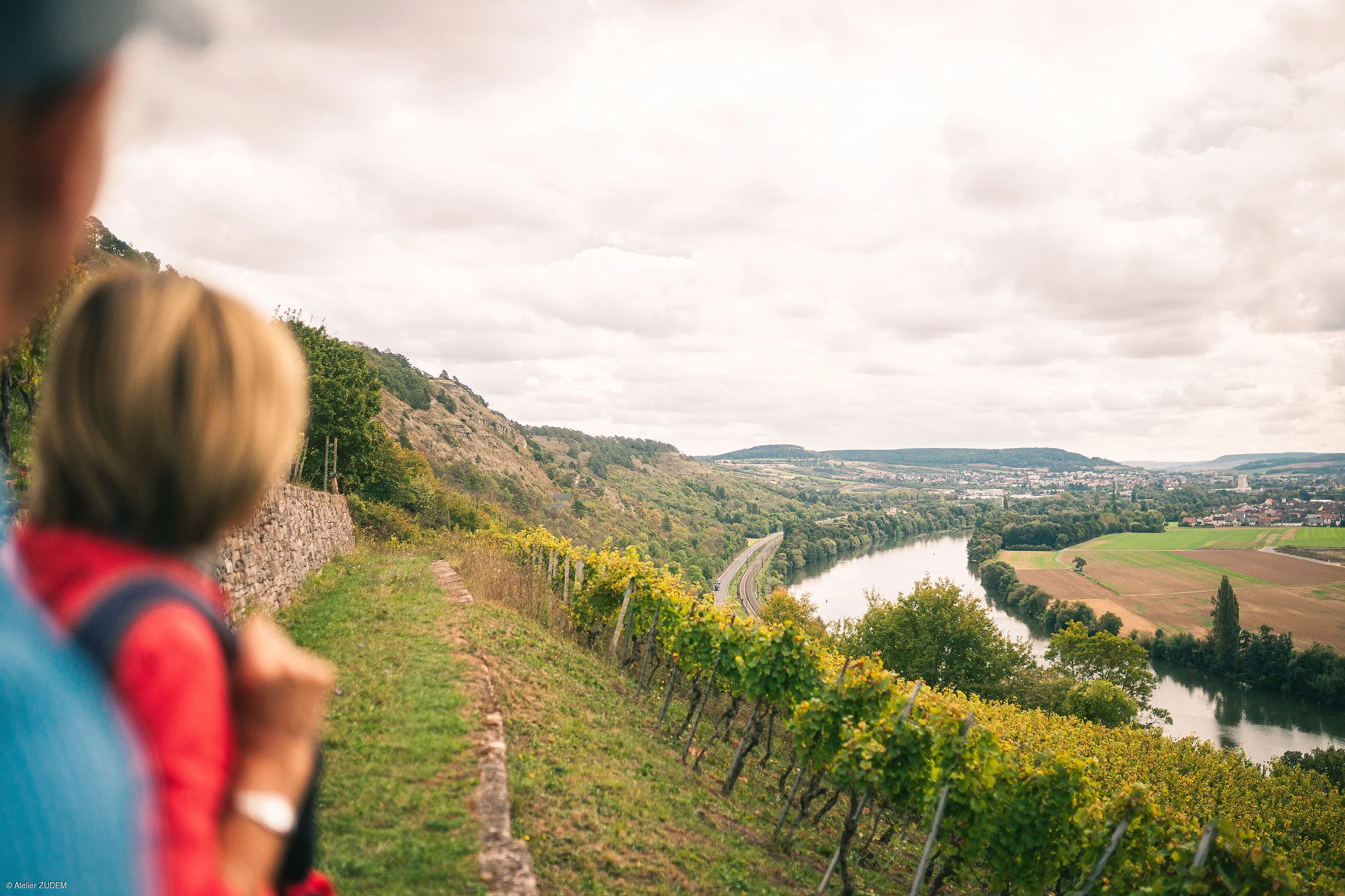 Blick über Weinberg mit Fluss und Feldern, unscharfe Personen im Vordergrund links sichtbar.