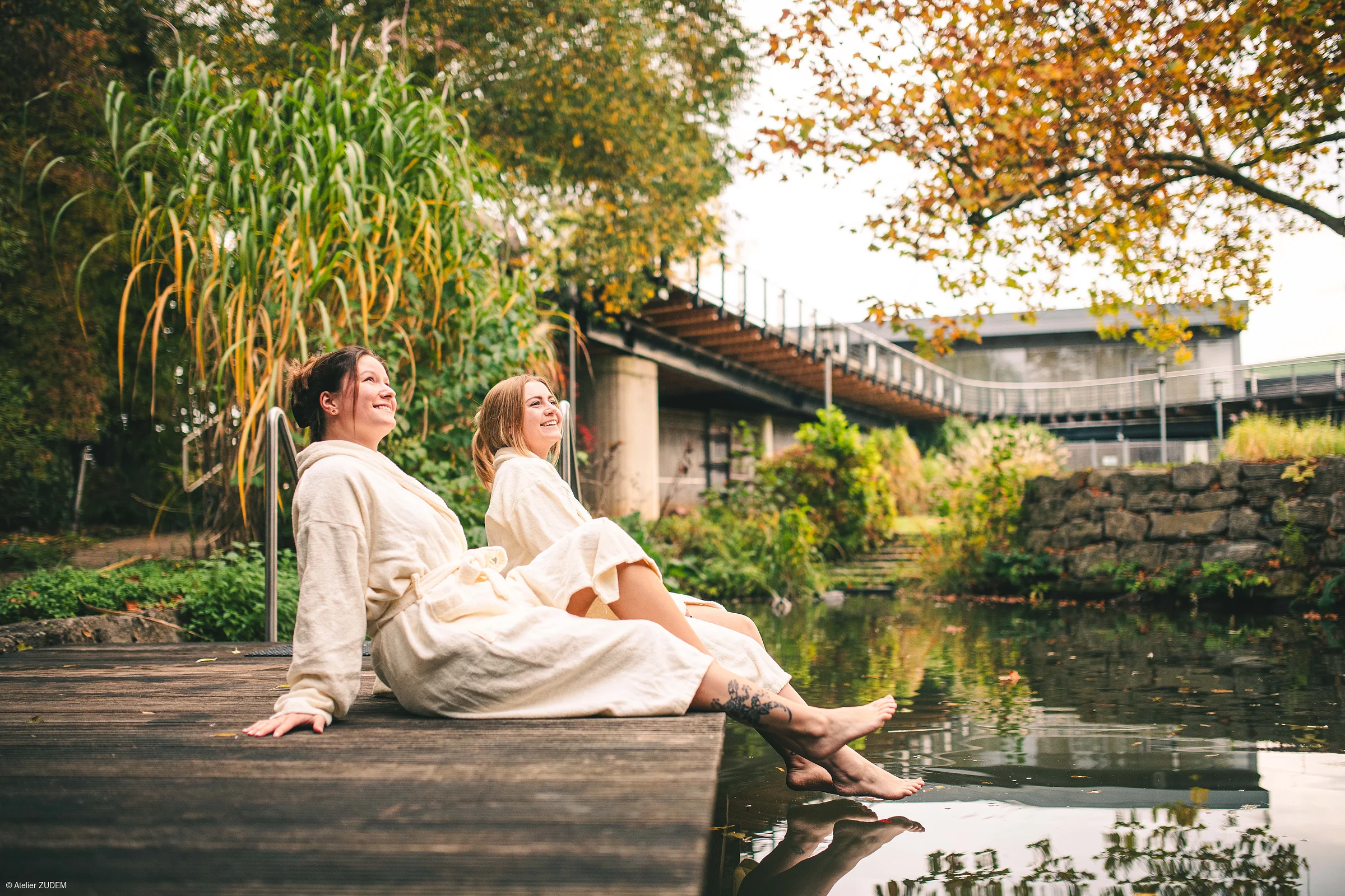 Zwei Frauen in Bademänteln sitzen am Holzsteg und halten ihre Füße ins Wasser eines Teichs im herbstlichen Park.