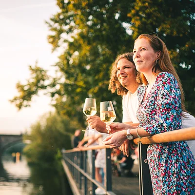 Zwei Personen lehnen an einem Geländer am Wasser und halten Weingläser bei Sonnenuntergang.