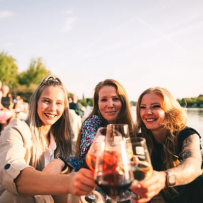 Drei Frauen sitzen am Wasser und stoßen mit Getränken in Gläsern an.