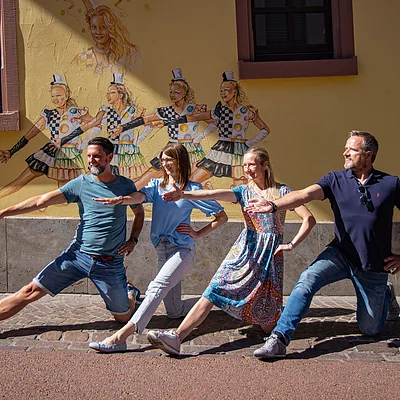 Vier Personen knien auf einem Gehweg vor einer Wand mit bunten Tanzfiguren und strecken jeweils einen Arm nach vorne aus.