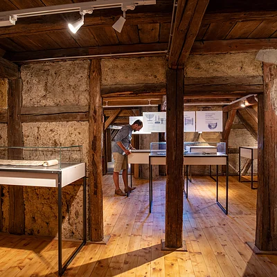 Museumsausstellung in einem Raum mit Holzbalken, Vitrinen und einer Person, die ein Exponat betrachtet