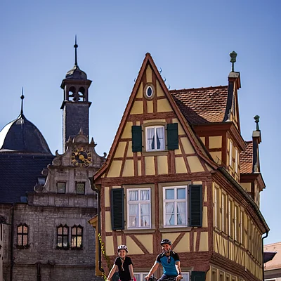 Zwei Radfahrer fahren vor einem Fachwerkhaus und einer historischen Uhrturmanlage bei klarem Himmel.