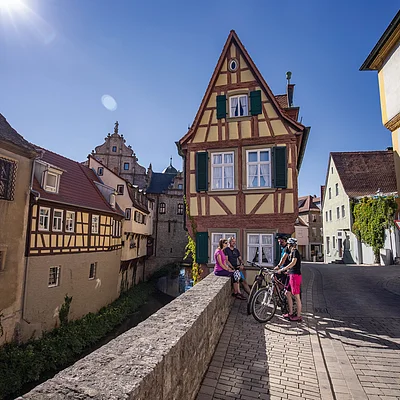 Vier Personen mit Fahrrädern stehen und sitzen an einer Brücke vor Fachwerkhäusern bei Sonnenschein.