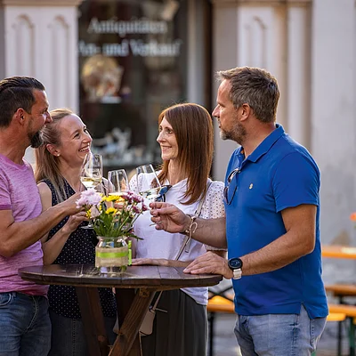 Vier Personen stehen an einem Stehtisch im Freien und stoßen mit Weingläsern an.