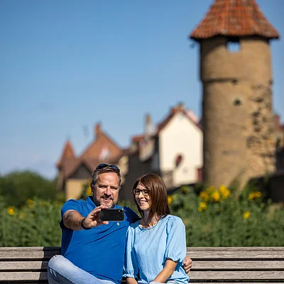 Paar sitzt auf Parkbank vor historischen Gebäuden und macht ein Selfie mit Smartphone.