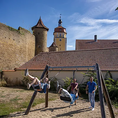 Zwei Personen schaukeln auf einem Spielplatz, zwei weitere stehen daneben, im Hintergrund historische Gebäude und blauer Himmel.