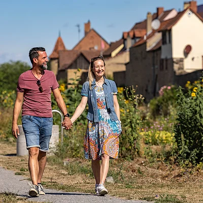 Paar hält Händchen und spaziert auf einem Weg mit Blumen und Häusern im Hintergrund bei Sonnenschein.