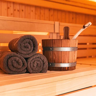 Drei braune Handtücher und ein Holzeimer mit Schöpfkelle auf einer Holzbank in einer Sauna.