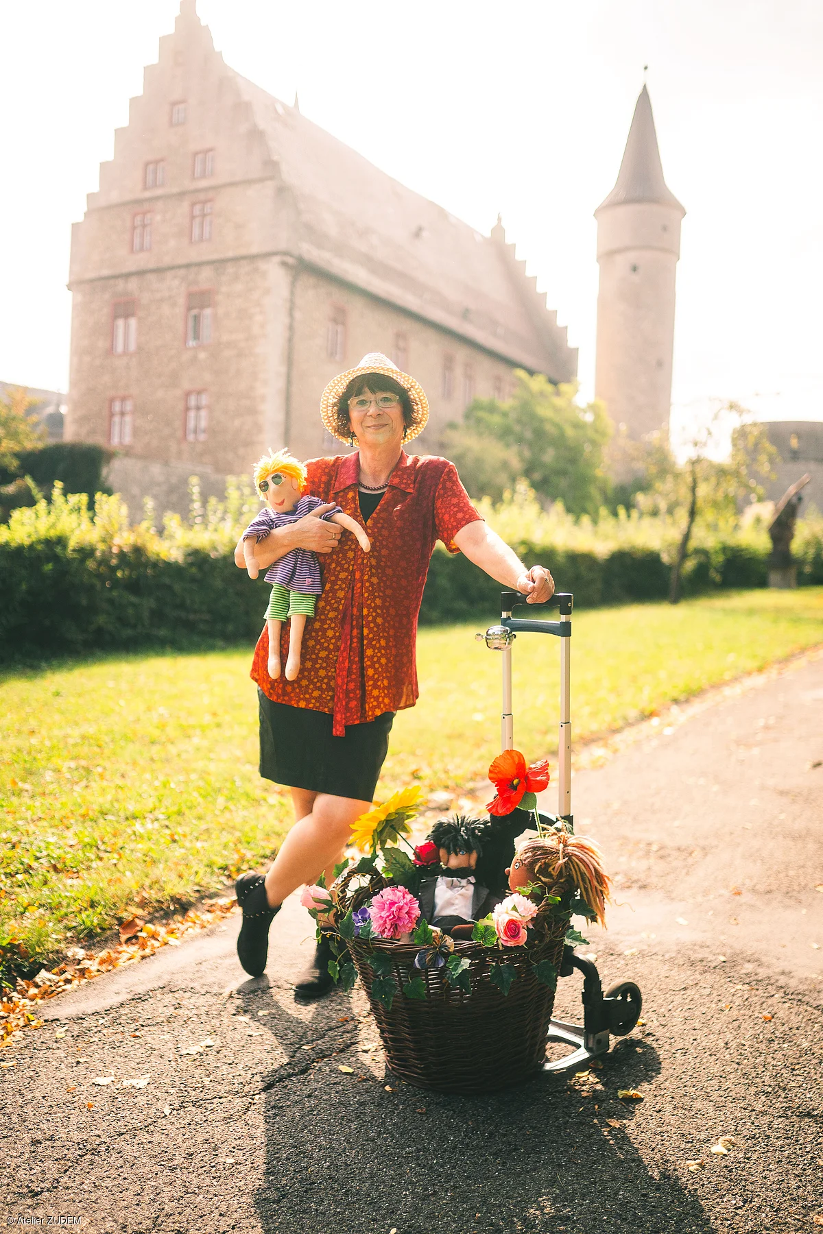 Frau mit Strohhut hält Puppe und steht neben Rollator mit Blumenkorb vor historischem Gebäude.