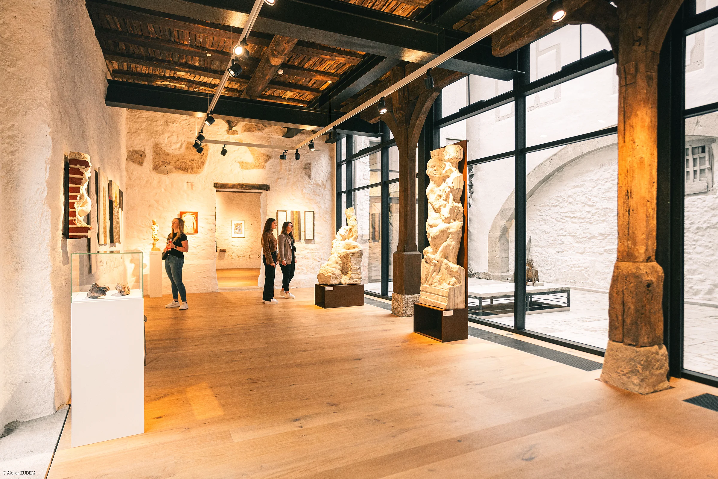 Innenraum eines Museums mit Holzdecke, großen Fenstern, Skulpturen und drei Personen, die Kunstwerke betrachten.