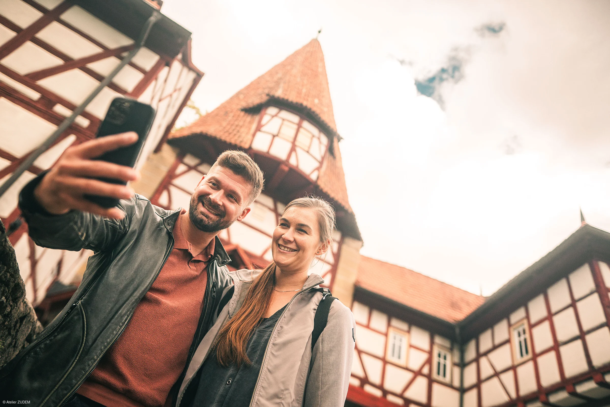 Zwei Personen machen vor einem Fachwerkhaus mit Turm ein Selfie mit dem Smartphone.