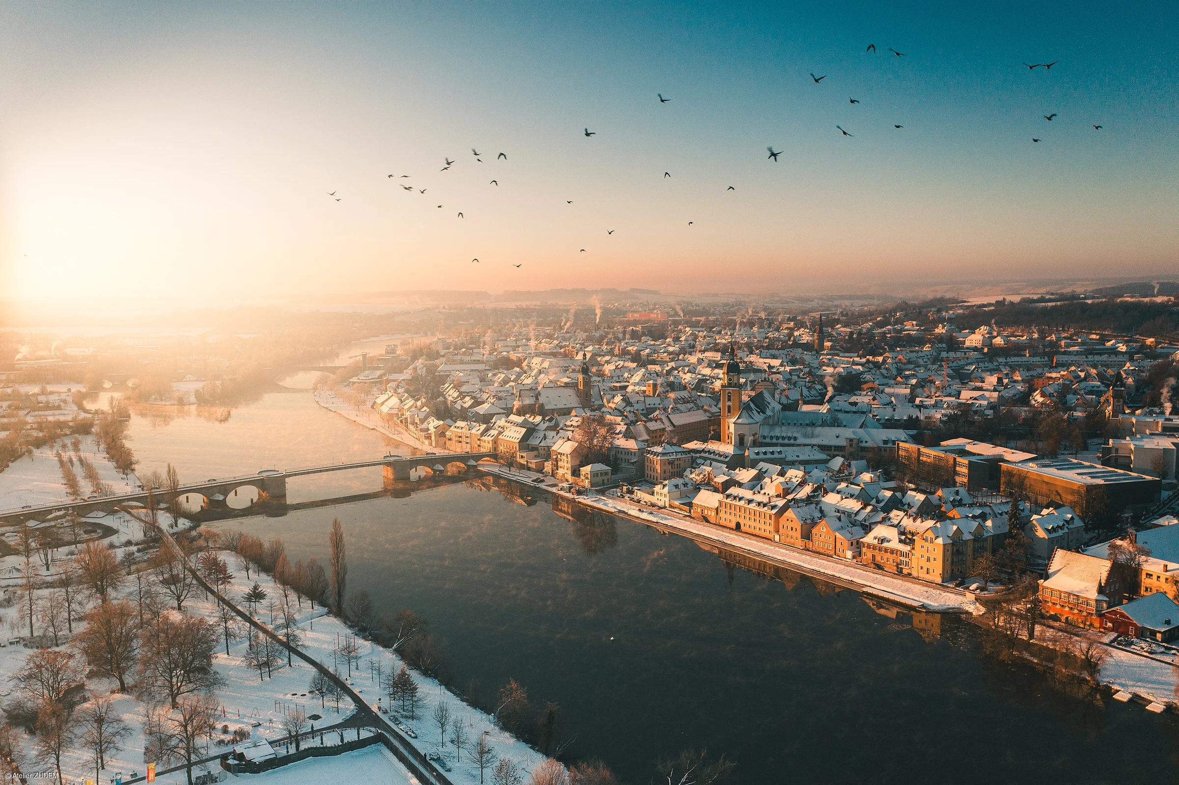 Vogelperspektive auf eine verschneite Stadt mit Fluss, Brücke und Vögeln im Sonnenaufgang.