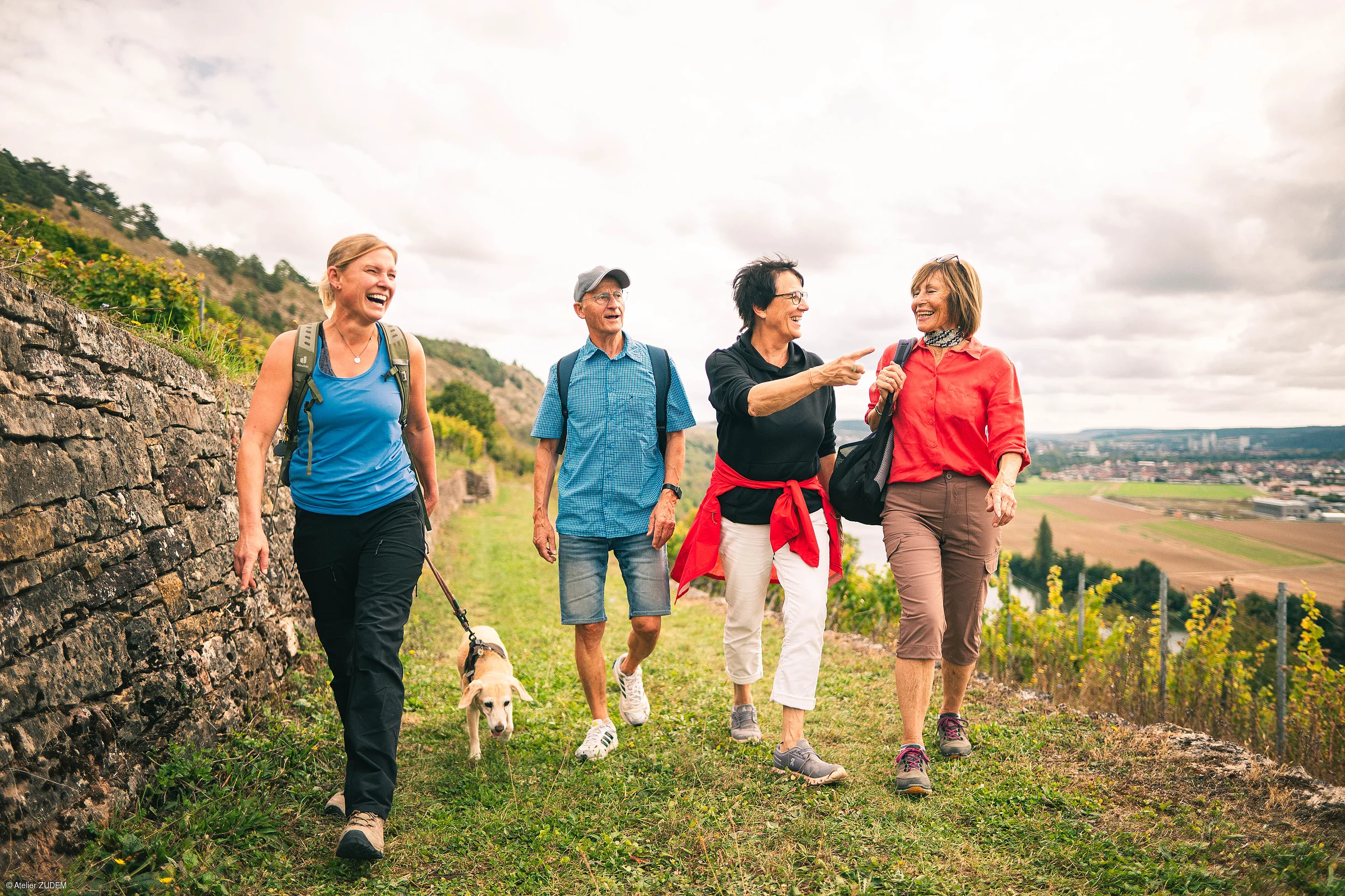 Vier Personen wandern auf einem Weg neben einer Steinmauer, eine Frau führt einen Hund an der Leine.