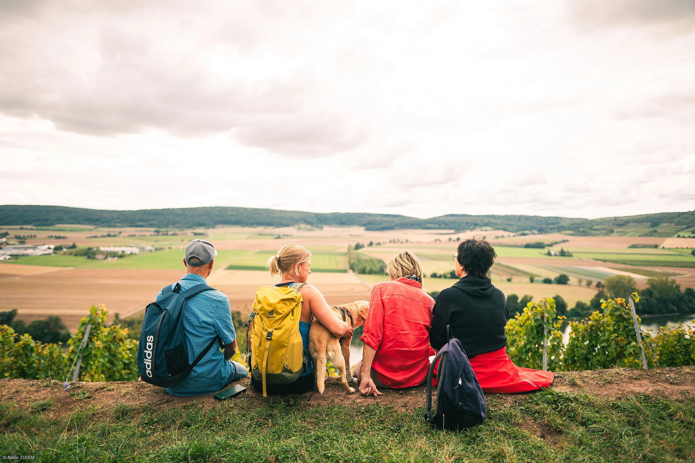 Vier Personen mit Rucksäcken und ein Hund sitzen auf einer Wiese mit Blick auf Felder und Hügel.