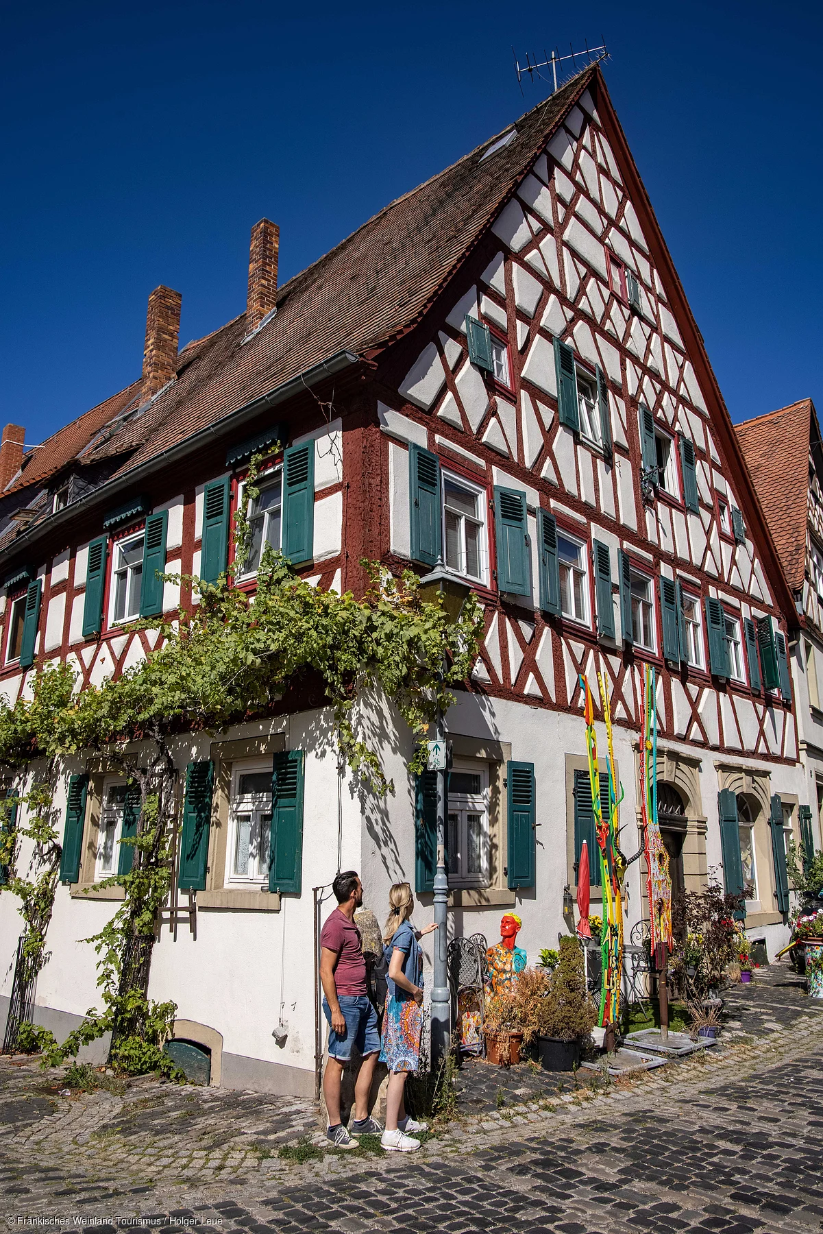 Fachwerkhaus mit grünen Fensterläden und zwei Personen auf Kopfsteinpflaster bei klarem Himmel.