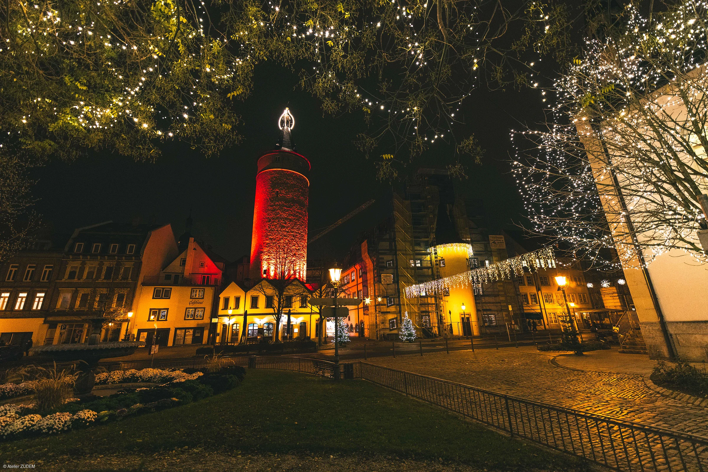 Nachtaufnahme eines beleuchteten Stadtplatzes mit altem Turm, Häusern und Weihnachtsbeleuchtung in Bäumen.