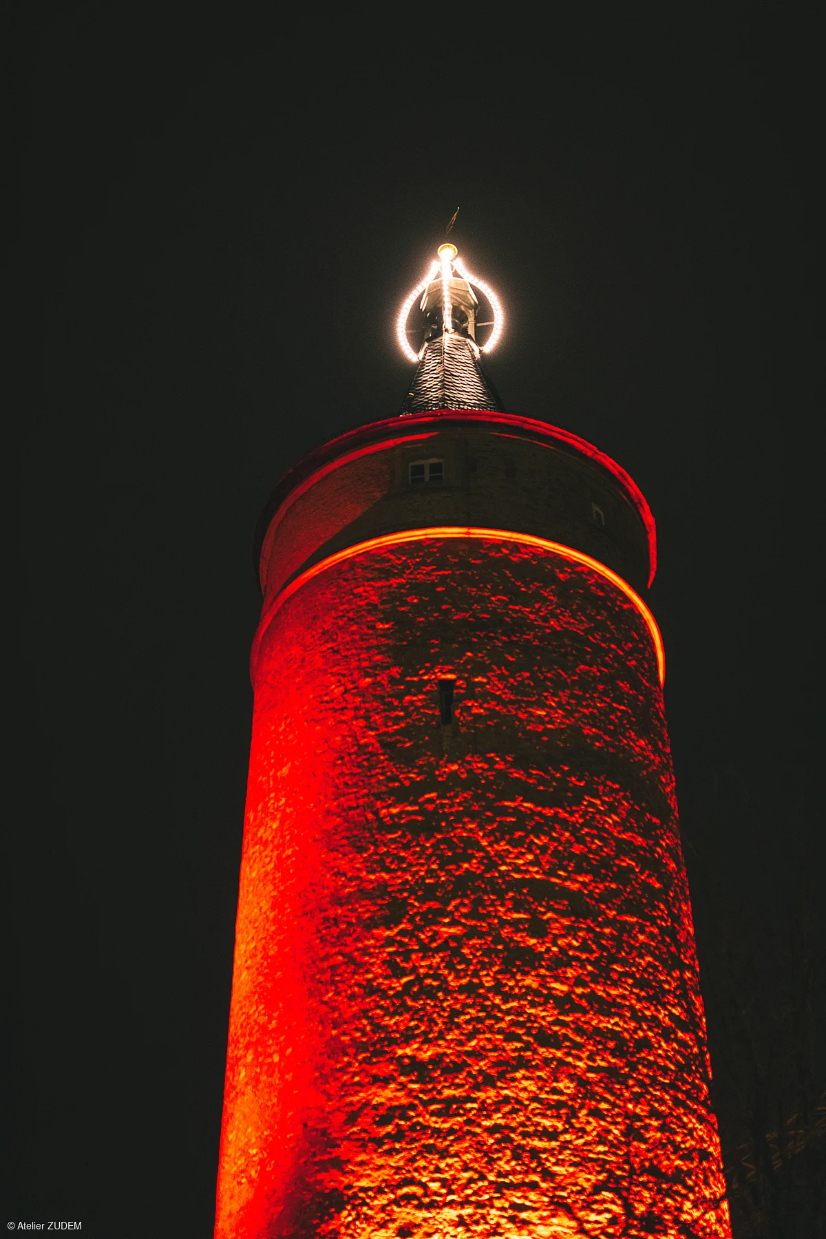 Turm mit roter Beleuchtung und leuchtendem Ring an der Spitze bei Nacht vor dunklem Himmel.