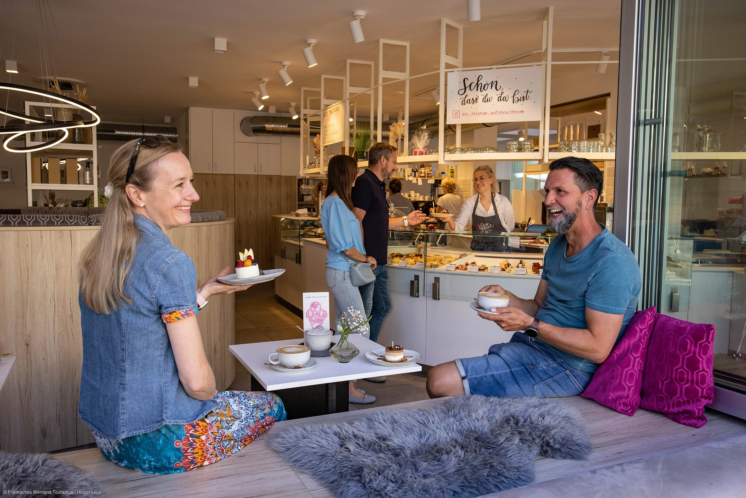 Zwei Personen sitzen mit Kaffee und Kuchen an einem Tisch in einem Café, im Hintergrund Bedienung und Kunden am Tresen.