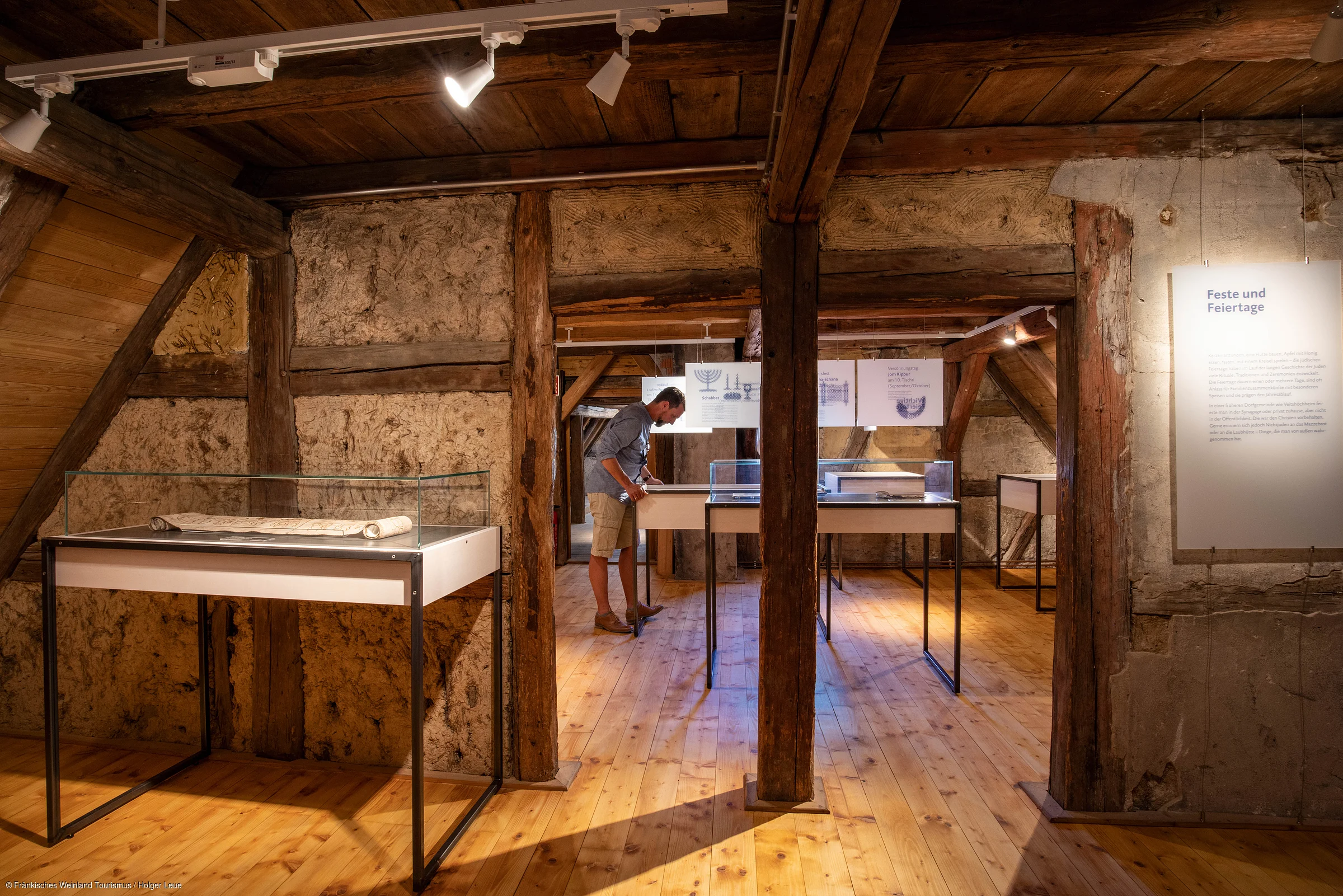 Museumsausstellung in einem Raum mit Holzbalken, Vitrinen und einer Person, die ein Exponat betrachtet