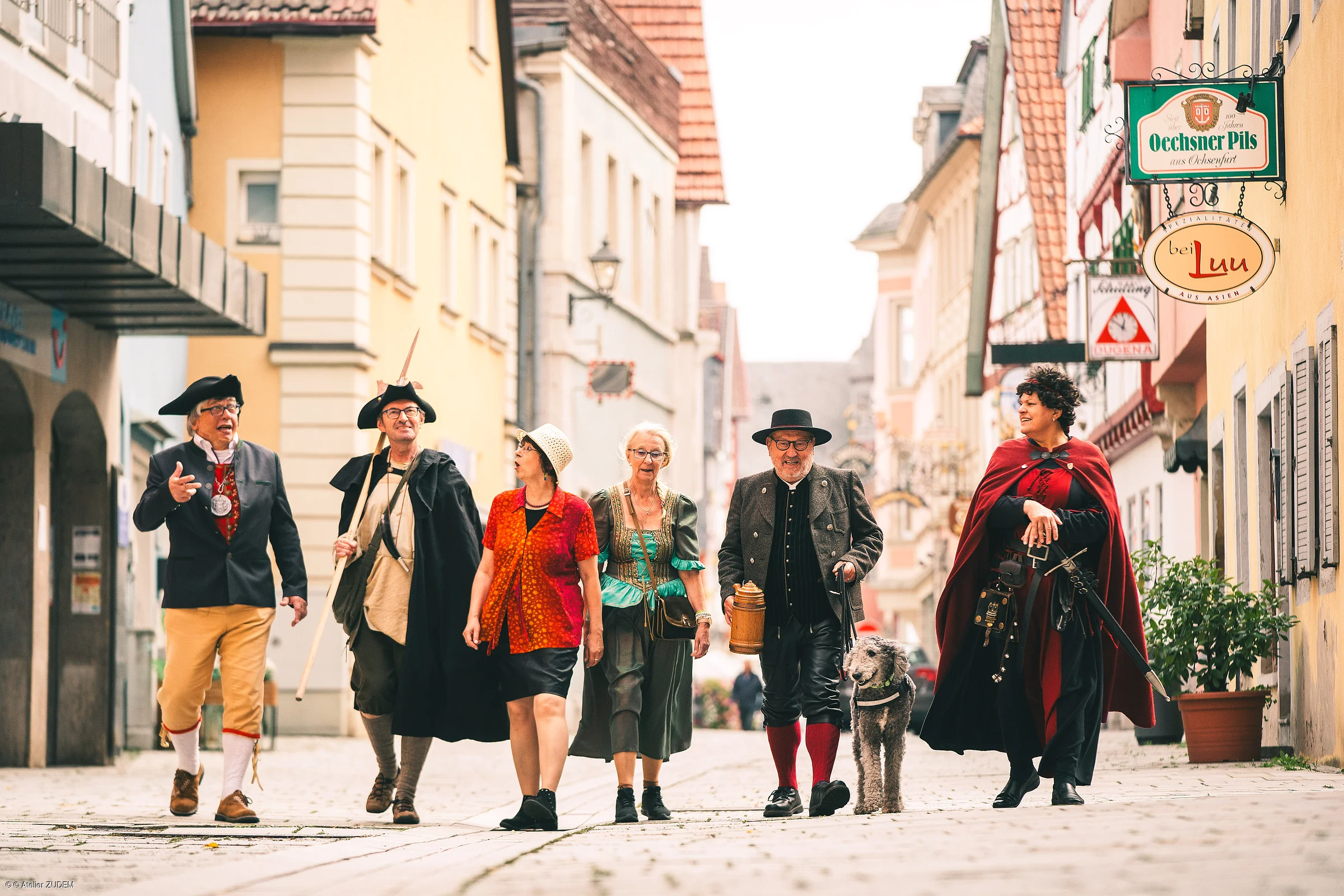 Sechs Personen in historischen Kostümen gehen eine gepflasterte Straße entlang, eine Person führt einen Hund an der Leine.