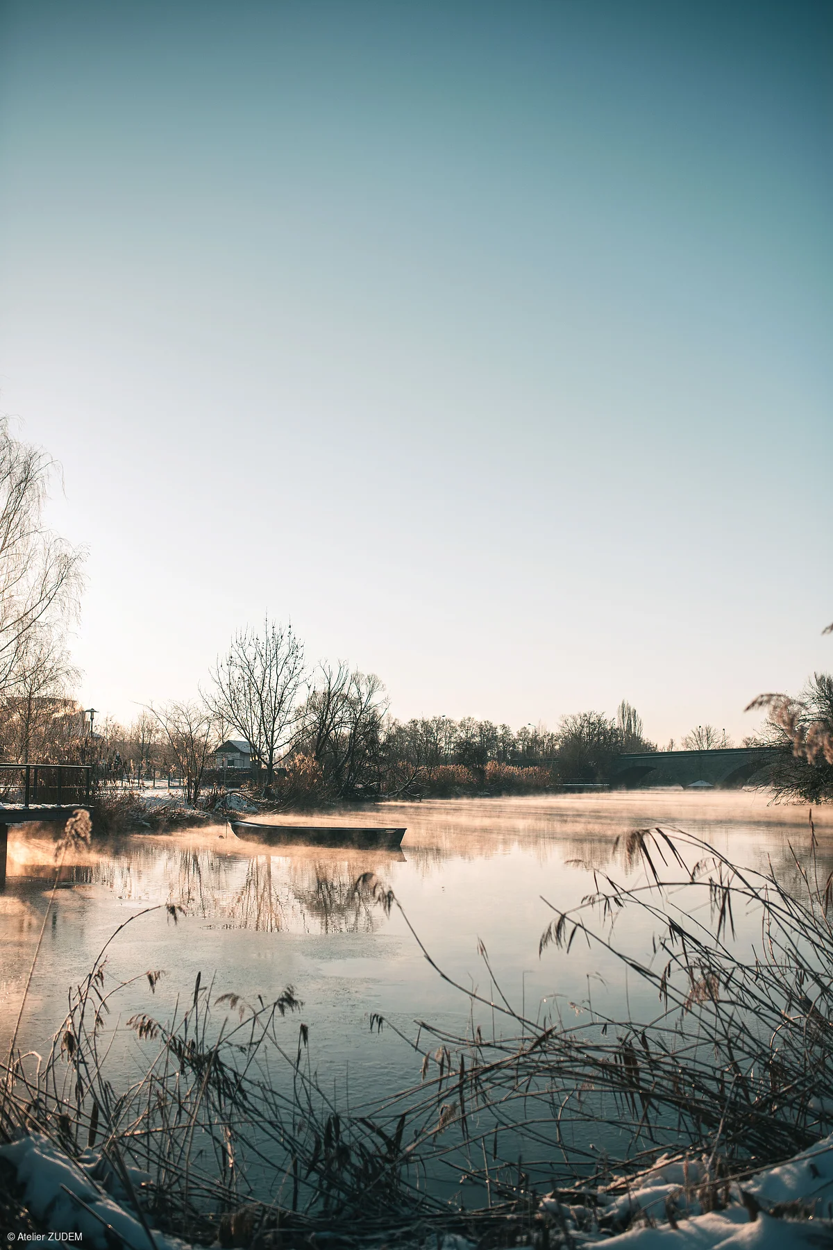 Fluss mit Nebel, kahlen Bäumen, Boot und Brücke bei klarem Himmel im Winter.
