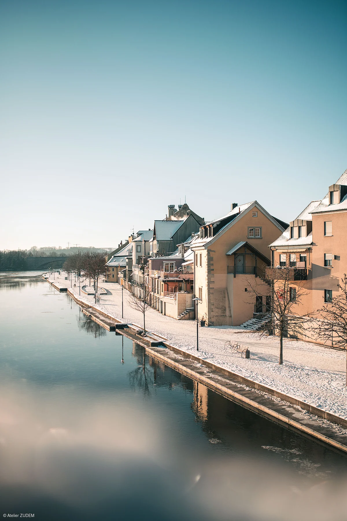 Schneebedeckte Häuser und Uferpromenade an einem ruhigen Fluss unter klarem Himmel im Winter.