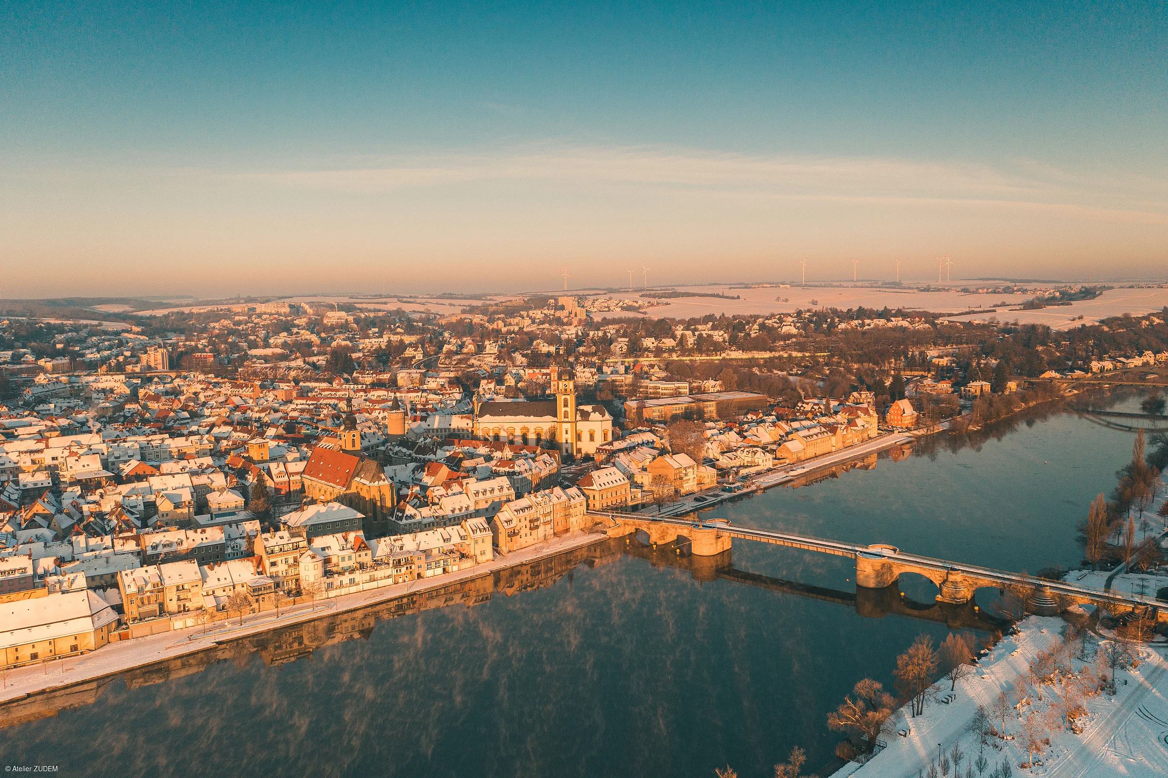 Luftaufnahme einer verschneiten Stadt mit Fluss und Brücke bei Sonnenaufgang oder Sonnenuntergang.