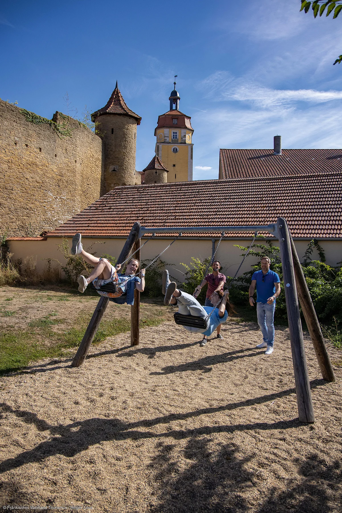 Zwei Personen schaukeln auf einem Spielplatz, zwei weitere stehen daneben, im Hintergrund historische Gebäude und blauer Himmel.