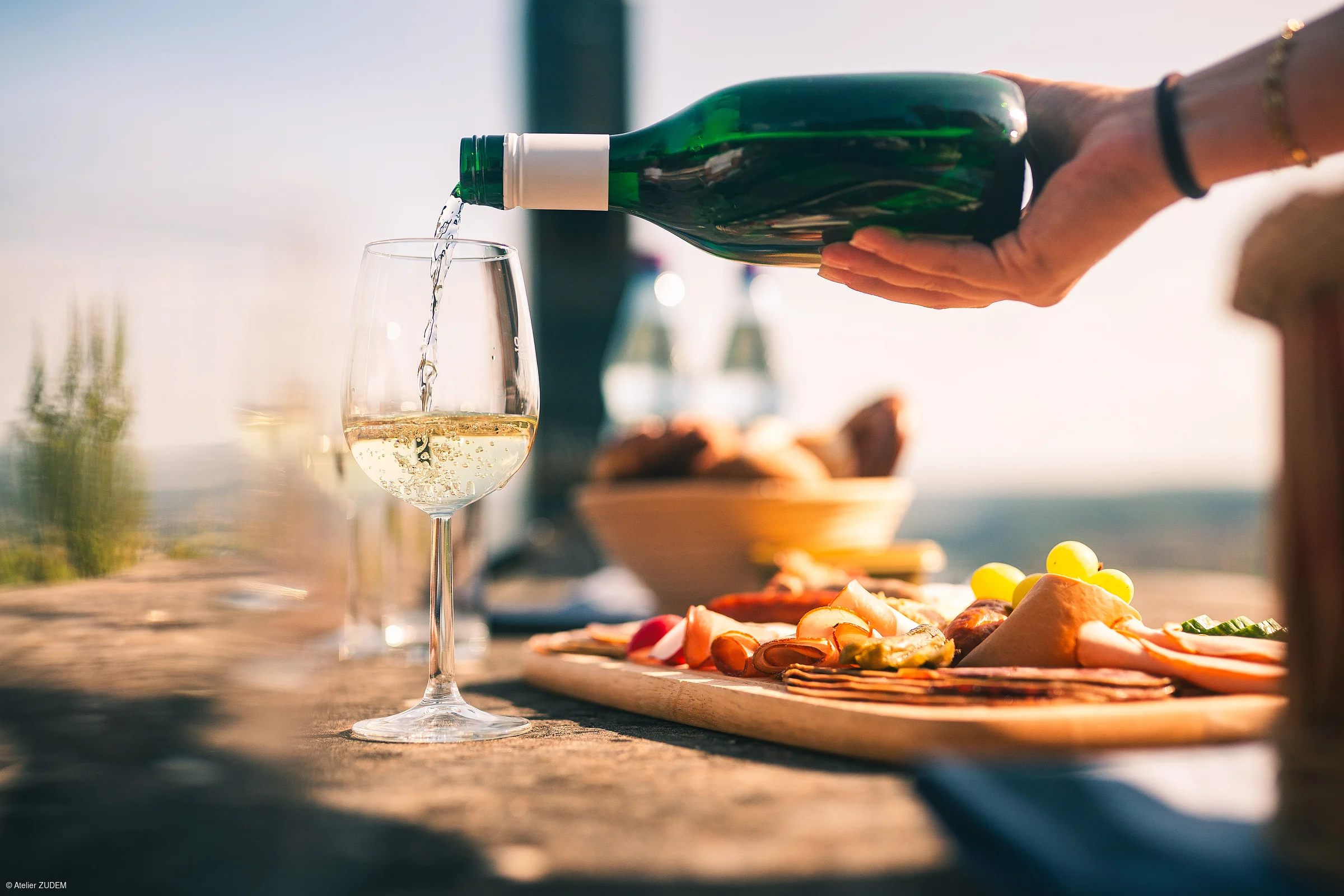 Weißwein wird aus einer grünen Flasche in ein Weinglas gegossen, daneben liegt eine Platte mit Aufschnitt und Trauben.