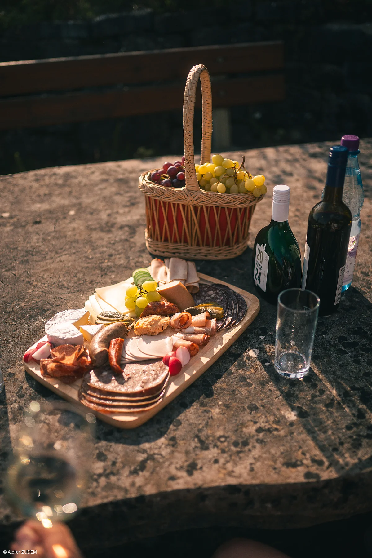 Brett mit verschiedenen Wurst- und Käsesorten, Traubenkorb, Weinflaschen und Glas auf Steintisch im Freien.