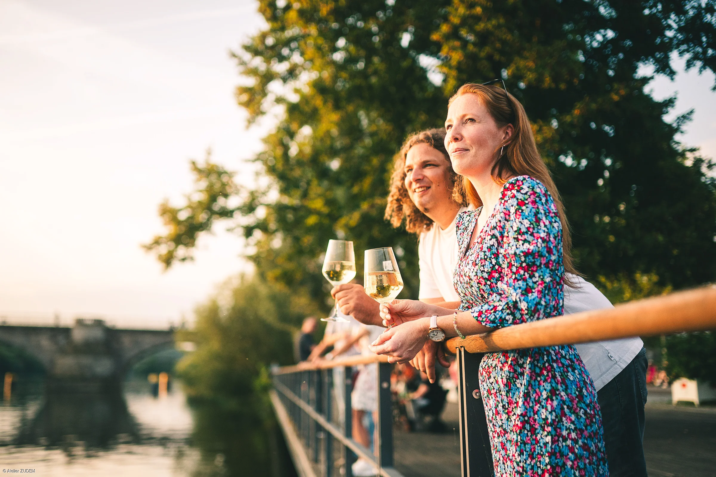 Zwei Personen lehnen an einem Geländer am Wasser und halten Weingläser bei Sonnenuntergang.