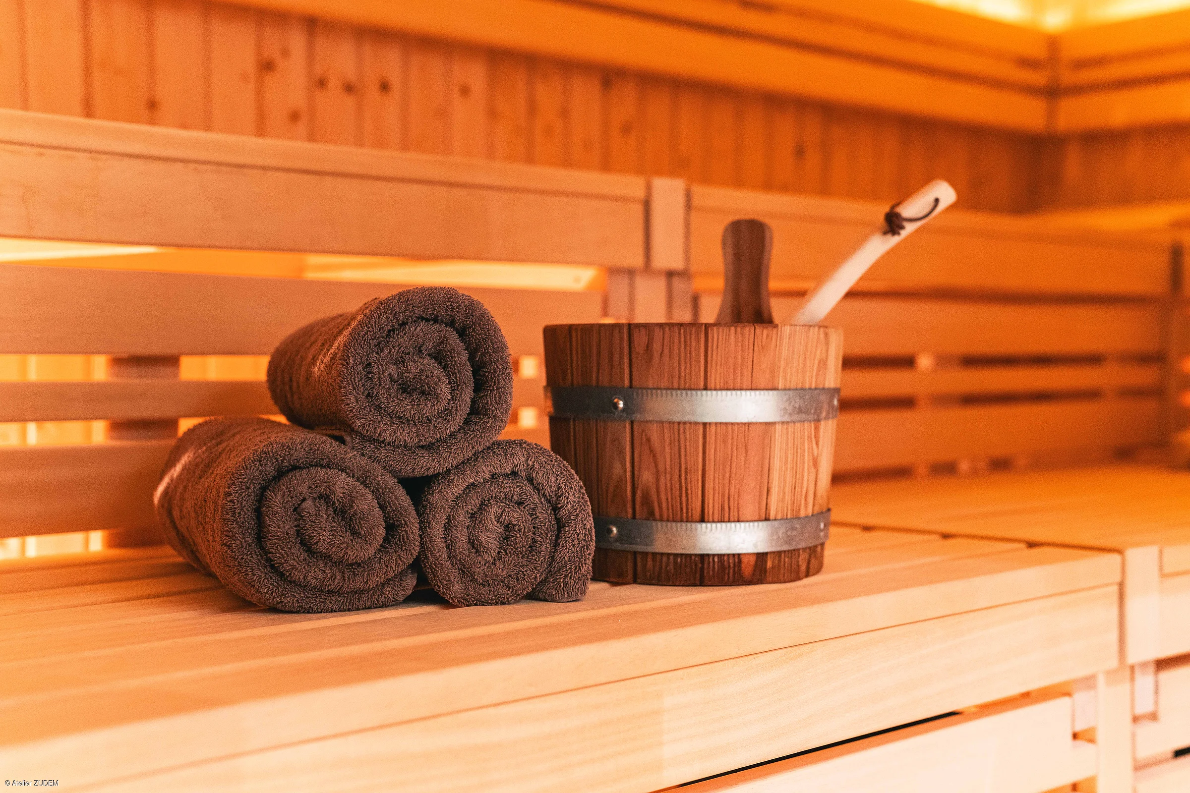 Drei braune Handtücher und ein Holzeimer mit Schöpfkelle auf einer Holzbank in einer Sauna.