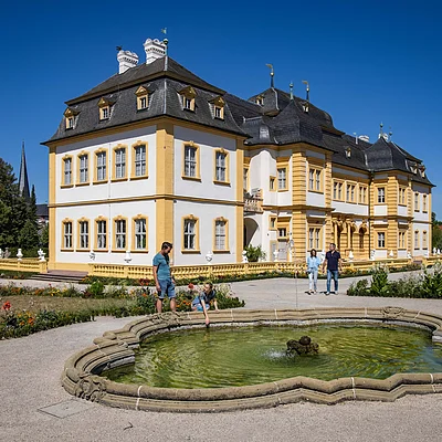 Barockschloss mit gelb-weißer Fassade, Brunnen im Vordergrund und vier Personen im Schlossgarten bei Sonnenschein