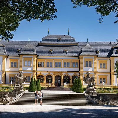 Barockschloss mit gelb-weißer Fassade, Treppe, Statuen und zwei Personen im Vordergrund bei blauem Himmel