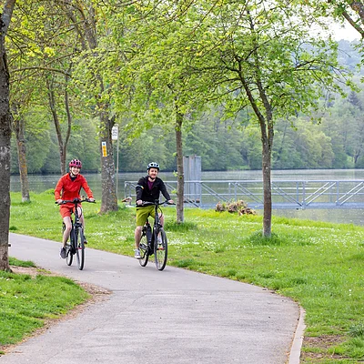 Zwei Personen fahren mit Fahrrädern auf einem Weg neben einem See, umgeben von Bäumen und Wiese.