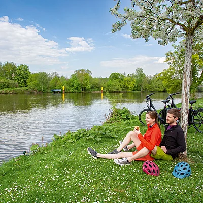Paar sitzt am Flussufer auf Gras unter blühendem Baum, zwei Fahrräder und Helme stehen daneben.