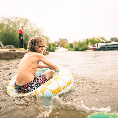 Kind sitzt in Schwimmring im Wasser, im Hintergrund Ufer mit stehender Person und Booten.