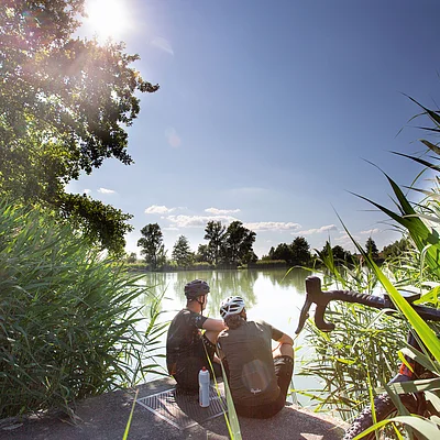 Zwei Radfahrer mit Helmen sitzen am Ufer eines Sees, umgeben von Schilf und Bäumen bei Sonnenschein.