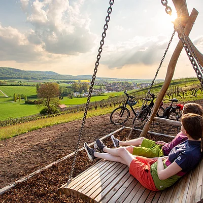 Zwei Personen sitzen auf einer Holzschaukel mit Blick auf grüne Felder und einen Ort im Hintergrund bei Sonnenlicht.
