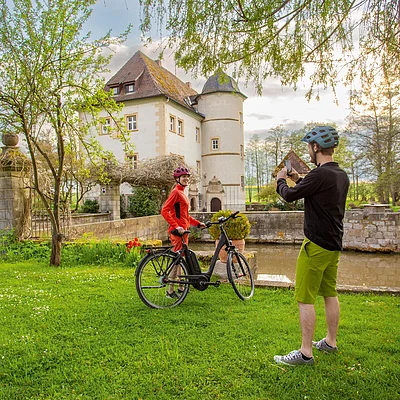 Zwei Personen mit Fahrradhelmen in einem Garten vor einem historischen Gebäude, eine macht ein Foto von der anderen.