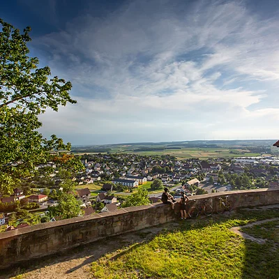 Blick von einer Anhöhe auf eine Stadt mit Häusern, zwei Personen an einer Mauer und einem Fachwerkhaus rechts.