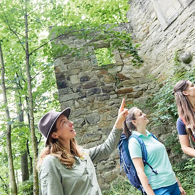 Drei Frauen mit Rucksäcken stehen vor einer alten Steinmauer im Wald, eine zeigt nach oben.
