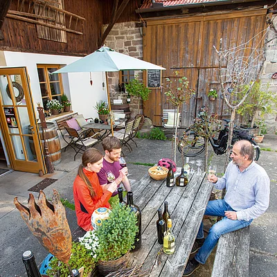 Drei Personen sitzen an einem Holztisch im Hof, trinken Wein, umgeben von Pflanzen und Weinflaschen.
