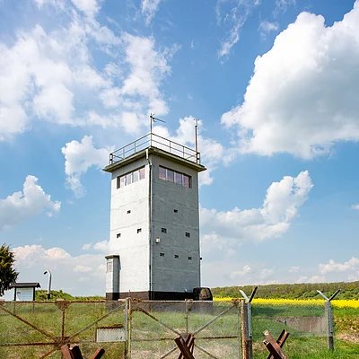 Alter grauer Wachturm hinter rostigem Maschendrahtzaun mit Panzersperren auf grünem Feld unter blauem Himmel.