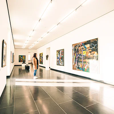 Frau betrachtet abstrakte Gemälde in einem hellen, modernen Kunstmuseum mit schwarzen Bodenfliesen.