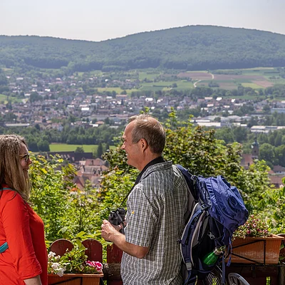 Zwei Wanderer mit Rucksäcken stehen auf einer Terrasse mit Blumen und blicken auf eine Stadt und bewaldete Hügel.