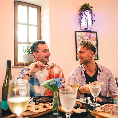 Zwei Männer sitzen an einem Tisch mit Essen und Getränken und unterhalten sich in einem hellen Raum.