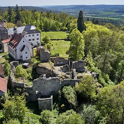 Luftaufnahme einer historischen Burganlage mit umliegenden Gebäuden und Wald in einer hügeligen Landschaft.