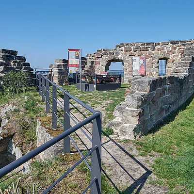 Ruine einer alten Steinmauer mit Infotafeln und Sitzgelegenheiten auf einer Anhöhe bei klarem Himmel.